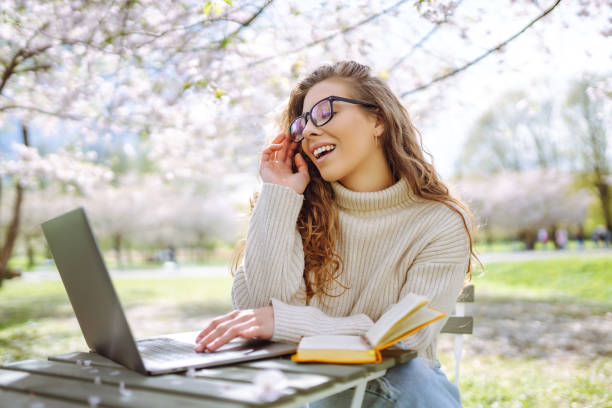 istock Young woman sitting on blooming park and using laptop. 2200134888