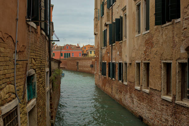 A narrow canal winds between old brick buildings with weathered facades. The green shutters of the historic buildings contrast with the warm hues of the houses. Traditional street in Venice, Italy.