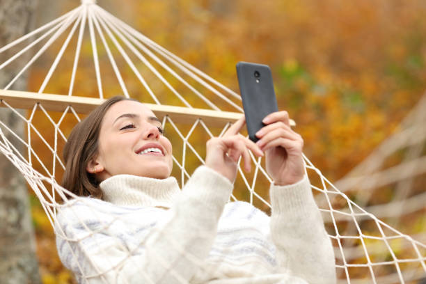 istock Happy woman using cell phone lying on hammock in autumn 2200160013