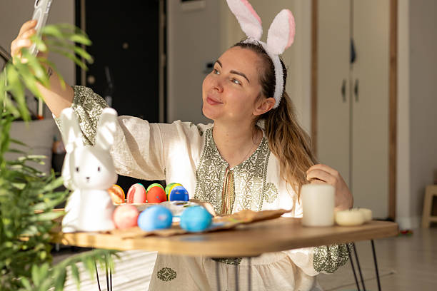 A smiling woman wearing bunny ears enjoys Easter festivities by taking a selfie while surrounded by vibrant eggs and decorations. Woman taking photo by mobile phone or having video call with family