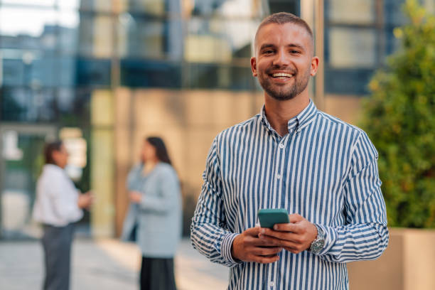 istock Young manager using smartphone smiling at camera in front of office building 2200182456