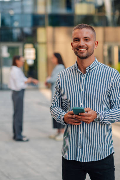 istock Young manager smiling and holding smartphone in front of office building 2200182465