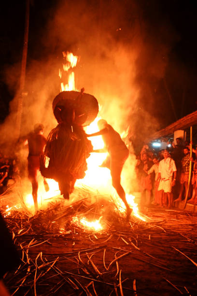 Thelasseri, Kannur, North Kerala, India - February 8, 2025. A Kudiveeran Thayyam dancer is captured mid-air, performing a dynamic and powerful leap over a large bonfire. The dancer wears traditional, colorful attire, complete with an elaborate headpiece, and is surrounded by a fiery glow as the flames crackle beneath them. The bonfire blazes brightly in the foreground, with orange and yellow flames flickering and creating a dramatic contrast against the night sky. The dancer’s body is positioned in a way that showcases both grace and strength, emphasizing the vibrant energy of the performance. The scene is set in an outdoor environment, with people gathered around, watching the captivating and intense ritual. The atmosphere is charged with excitement and reverence.