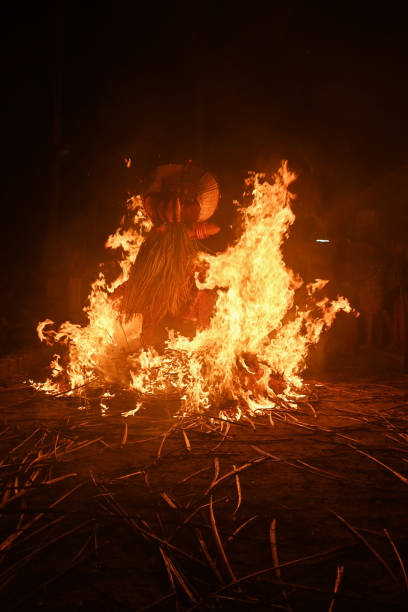 Thelasseri, Kannur, North Kerala, India - February 8, 2025. A Kudiveeran Thayyam dancer is captured mid-air, performing a dynamic and powerful leap over a large bonfire. The dancer wears traditional, colorful attire, complete with an elaborate headpiece, and is surrounded by a fiery glow as the flames crackle beneath them. The bonfire blazes brightly in the foreground, with orange and yellow flames flickering and creating a dramatic contrast against the night sky. The dancer’s body is positioned in a way that showcases both grace and strength, emphasizing the vibrant energy of the performance. The scene is set in an outdoor environment, with people gathered around, watching the captivating and intense ritual. The atmosphere is charged with excitement and reverence.
