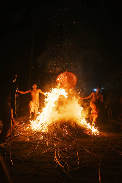 Thelasseri, Kannur, North Kerala, India - February 8, 2025. A Kudiveeran Thayyam dancer is captured mid-air, performing a dynamic and powerful leap over a large bonfire. The dancer wears traditional, colorful attire, complete with an elaborate headpiece, and is surrounded by a fiery glow as the flames crackle beneath them. The bonfire blazes brightly in the foreground, with orange and yellow flames flickering and creating a dramatic contrast against the night sky. The dancer’s body is positioned in a way that showcases both grace and strength, emphasizing the vibrant energy of the performance. The scene is set in an outdoor environment, with people gathered around, watching the captivating and intense ritual. The atmosphere is charged with excitement and reverence.