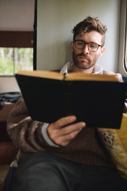 istock Man, reading and relax with book at home for novel study, literature learning and history information. Male person, caravan and glasses with storytelling for knowledge, philosophy journal and fiction 2200187565