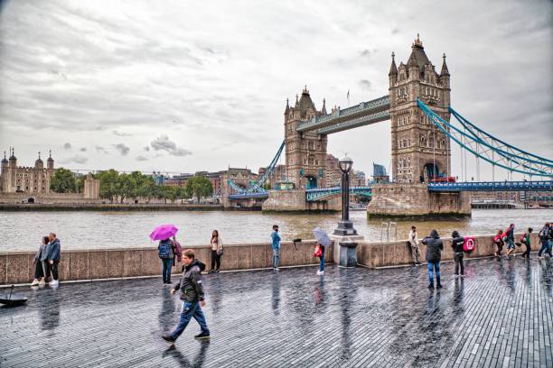 Tourists visit The Queen's Walk along river Thames in London with famous Tower Bridge in rainy weather.