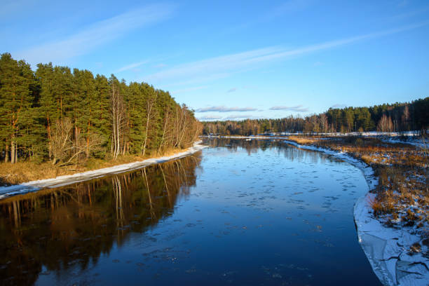 istock A partially frozen river winding through a winter landscape, surrounded by trees and snow-covered banks under a clear blue sky. 2200202581