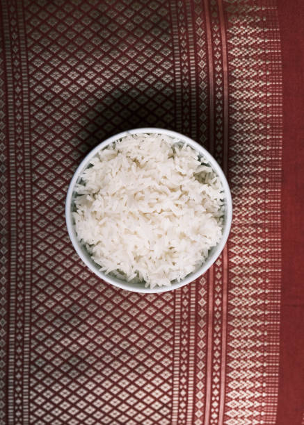 This image showcases a bowl of steamed jasmine rice, a staple of Thai cuisine, elegantly presented in a traditional Thai ceramic bowl. The bowl is placed on a richly patterned red fabric, enhancing the cultural and authentic feel of the composition. The fluffy texture and delicate aroma of jasmine rice make it a perfect pairing for a variety of Thai dishes. Ideal for use in restaurant branding, menu designs, or food photography, this image highlights the simplicity and importance of this essential Thai ingredient.