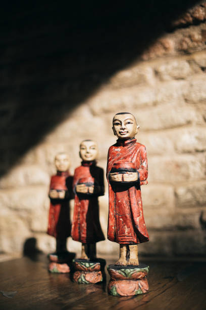 This image features a row of traditional Thai monk statues, each holding an alms bowl, symbolizing Buddhist spirituality and the practice of giving. The statues are adorned in red robes with intricate detailing and are displayed against a rustic brick wall, with warm sunlight casting soft shadows. This composition creates a serene and culturally rich atmosphere, making it ideal for Thai restaurant interior design, branding, or cultural photography that highlights Buddhist traditions.