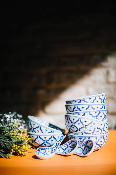 A beautifully arranged stack of traditional Thai ceramic bowls with intricate blue and white floral patterns, placed on a wooden surface with soft sunlight illuminating the scene. The rustic brick wall in the background adds an authentic and warm ambiance, enhancing the cultural and artistic essence of Thai craftsmanship. These handcrafted bowls are commonly used in Thai restaurants for serving soups, curries, and other traditional dishes. The delicate floral motifs and fine ceramic work reflect the rich artistic heritage of Thailand, making this image ideal for themes related to Asian cuisine, restaurant decor, and cultural authenticity.