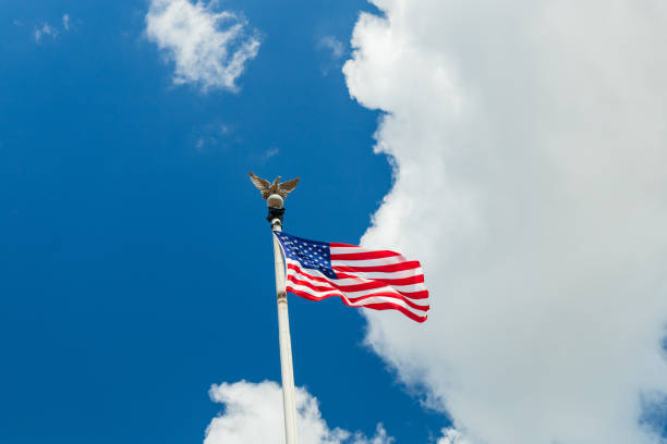 A large American flag is flying high in the sky. The sky is blue with a few clouds scattered throughout. The eagle on the flag is perched on a pole, and the flag is waving in the breeze