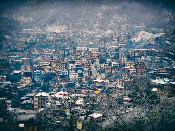 The breathtaking view of a hillside town features colorful multi-story buildings cascading down the slopes. A light layer of snow blankets the rooftops and trees, adding charm to the landscape. Misty mountains in the background create a picturesque contrast between nature and urban life of Solan, Himachal Pradesh. This image highlights the unique Lomo photography/Lomography style.
