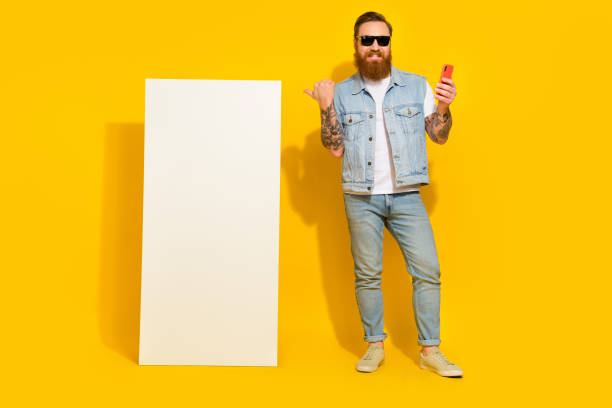 istock Photo of funny charming guy dressed denim vest sunglass pointing thumb modern gadget empty space isolated yellow color background 2200227581