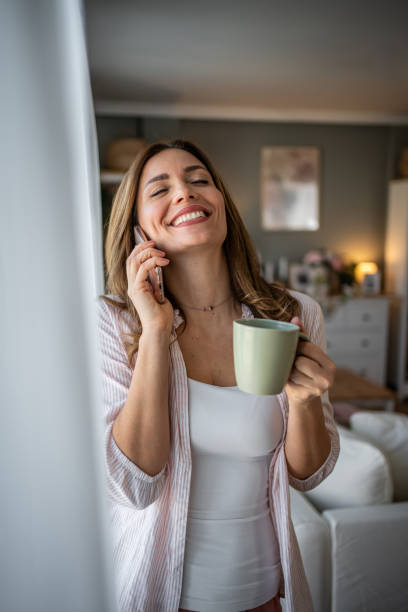 istock A young woman is smiling while holding a green cup and using her mobile phone, radiating happiness and relaxation in a cozy home environment 2200229287