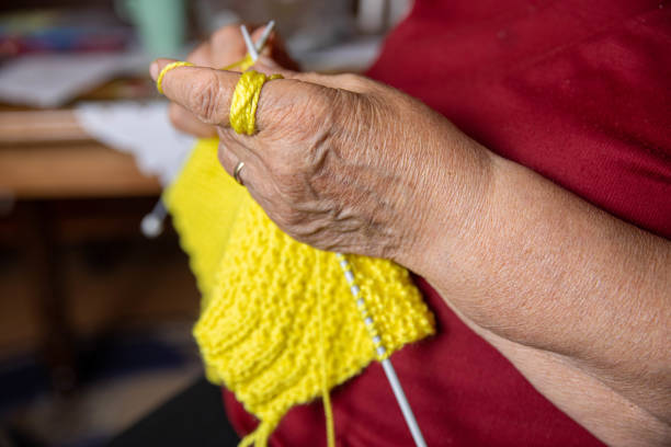 Portrait of an elderly woman in casual clothes, wearing glasses and knitting a yellow scarf for her daughter. Close up.