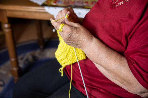 Portrait of an elderly woman in casual clothes, wearing glasses and knitting a yellow scarf for her daughter. Close up.