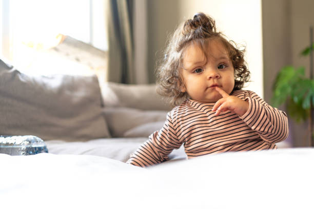 A curly-haired baby girl in a striped long-sleeve top sits on a soft couch, touching her lips with her finger in a thoughtful pose. Sunlight streams in through the window, illuminating her curious expression. The cozy home environment and natural light create a warm and peaceful scene of early childhood wonder.