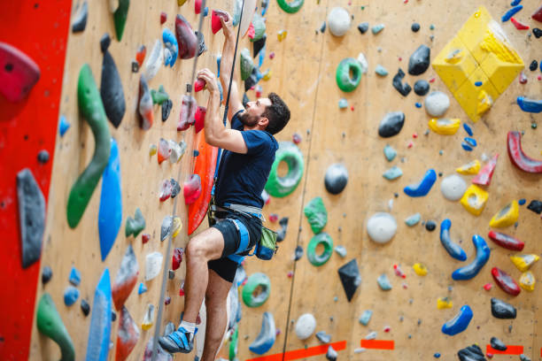 An adult man focuses on rock climbing at an indoor gym, showcasing agility and strength on a colorful climbing wall. Embracing an active lifestyle, he navigates the challenge with effort and determination.