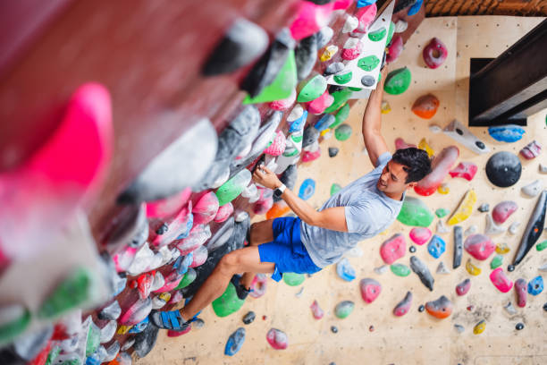 A young adult male athlete bouldering on a vibrant indoor climbing wall. He demonstrates agility and strength as he moves upward. The colorful holds add to the challenge of the exercise, showcasing an active and healthy lifestyle.