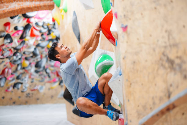 A young adult male athlete skillfully climbing a colorful indoor bouldering wall. Engaging in a challenging workout, he exhibits strength and agility, showcasing his sport climbing expertise in a vibrant rock climbing gym.