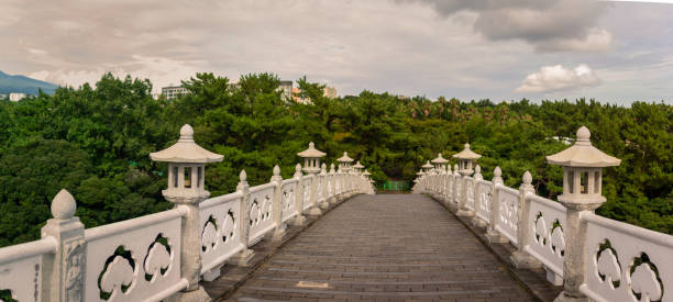 Scenic arched stone bridge adorned with white railings and traditional lanterns stretching into lush greenery under a cloudy sky on Jeju Island offering a tranquil atmosphere. Captured in Jeju, South Korea, Asia