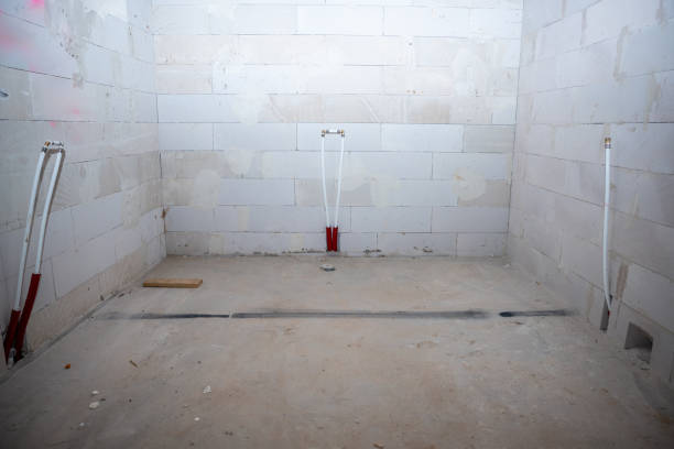 Interior under construction with white block walls, bare concrete floor, visible plumbing pipes, and a wooden plank on the ground.