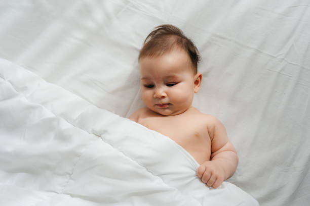 A happy baby lying on a soft bed with a white duvet. The baby is smiling and looks cheerful. The room is bright and sunny, with sunlight coming through the window, making everything feel warm and peaceful.