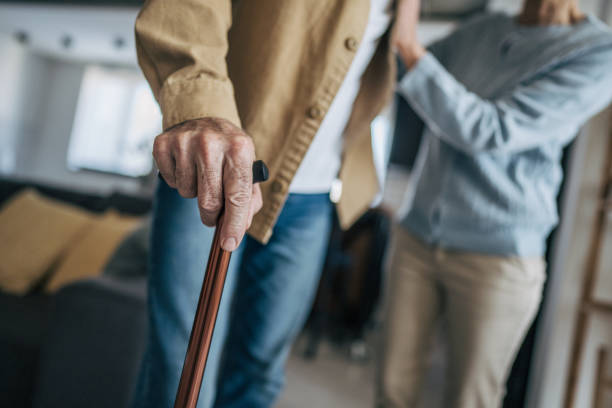 Close-up of a wrinkled hand gripping a walking cane, with a blurred figure of a nurse providing support in the background, highlighting the challenges and care associated with aging