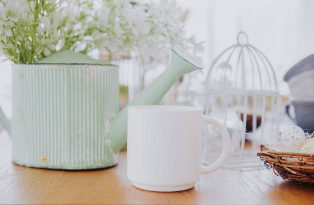 A white mug sits on a wooden table next to a green watering can and decor.