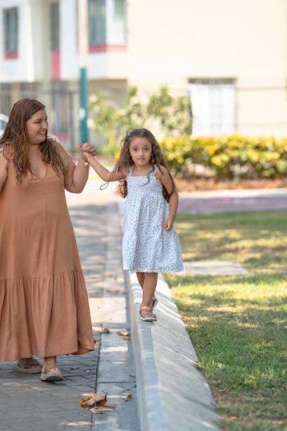 Preschool-aged girl carefully balances on a curb while holding her mother's hand, enjoying a sunny day at the park