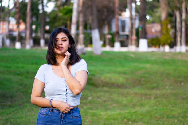 Beautiful girl in the park, she is standing over a grassy area, wearing a light blue shirt, smiling at camera