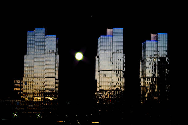 A striking nighttime cityscape featuring three modern glass skyscrapers illuminated by ambient light. The reflective surfaces capture the warm hues of the fading sunset and the glow of urban lights, creating a mesmerizing contrast against the dark night sky. A luminous full moon adds an ethereal touch to the composition, enhancing the futuristic and surreal aesthetic.