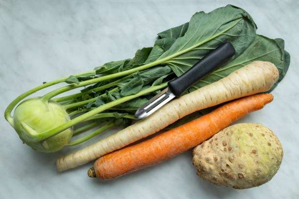Fresh kohlrabi, carrot, parsley root, and celery are laid out on a marble surface, accompanied by a vegetable peeler. The peeler rests next to the vegetables, highlighting their vibrant colors and textures against the smooth, elegant backdrop.