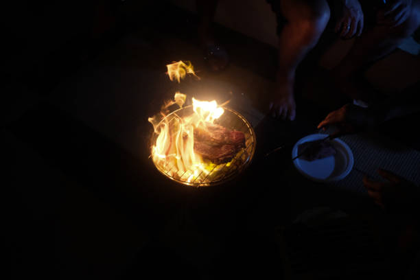 A dramatic scene of an open-flame barbecue at night, with steaks grilling over a small charcoal grill. Bright flames rise around the meat, illuminating the surrounding hands and plates in a dimly lit setting.