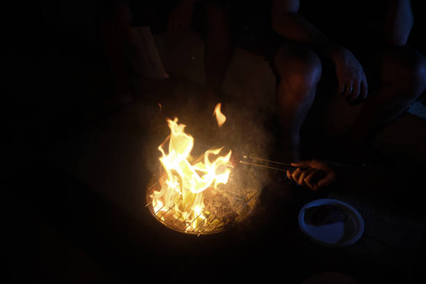 A dramatic scene of an open-flame barbecue at night, with steaks grilling over a small charcoal grill. Bright flames rise around the meat, illuminating the surrounding hands and plates in a dimly lit setting.