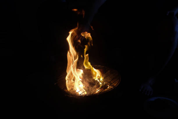 A dramatic scene of an open-flame barbecue at night, with steaks grilling over a small charcoal grill. Bright flames rise around the meat, illuminating the surrounding hands and plates in a dimly lit setting.