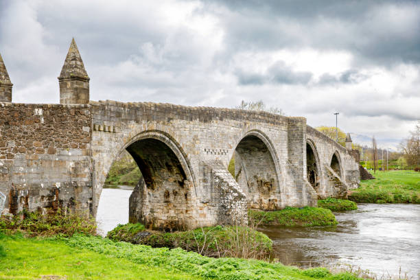 istock Historic stone bridge over a flowing river in Stirling, Scotland, surrounded by lush greenery and dramatic cloudy skies 2200335233