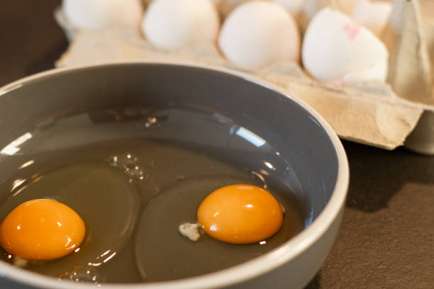 Close up of two cracked eggs in a bowl on grey kitchen table with dozen of white eggs in a cardboard