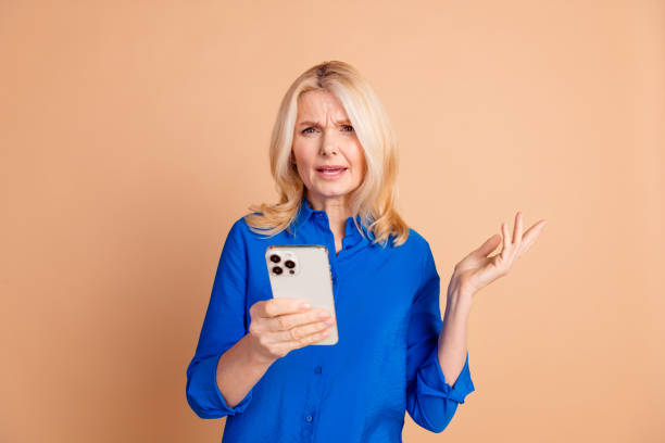 istock Stylish Senior Woman Wearing a Blue Shirt Expressing Confusion While Using Smartphone Against Beige Background 2200341039