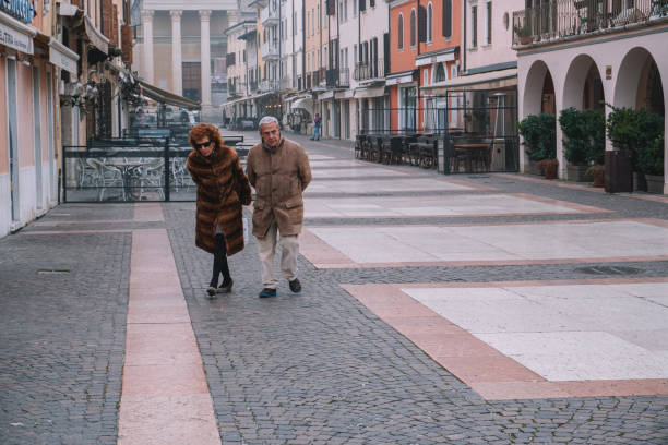 Bordolino, Italy - 06.02.2024. An elderly couple walks arm in arm down a quiet, picturesque Italian street. The cobblestone pathway and pastel-colored buildings create a charming, timeless atmosphere.