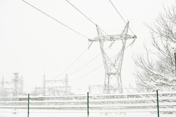 A large electrical pylon and power lines covered in snow stand against a winter sky, with an industrial substation visible behind a fence in cold weather conditions.