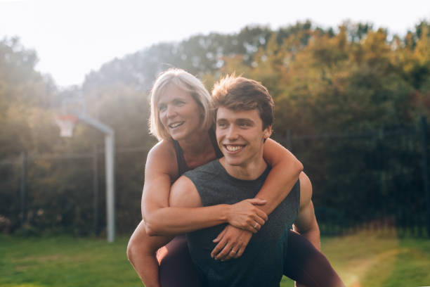 A smiling mother gets a piggyback ride from her son in a sunny outdoor setting, enjoying quality time and bonding together