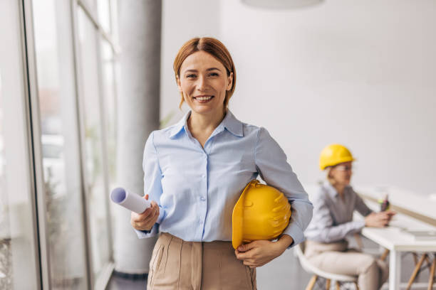 istock Female architect holding blueprint and hardhat smiling at camera in office 2200368967