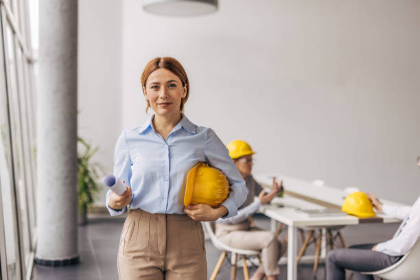 istock Confident female architect holding plans and hard hat in office 2200369013