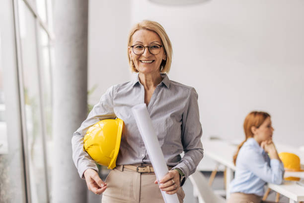 istock Senior female architect holding blueprint and hardhat smiling at camera 2200369214