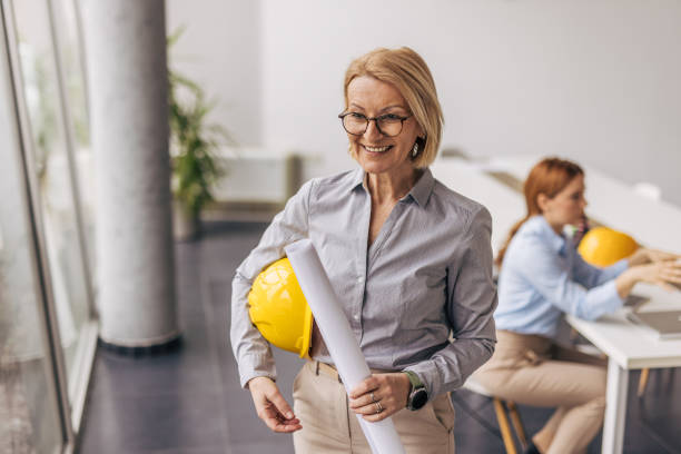 istock Smiling female architect holding blueprint and hardhat in office 2200369215