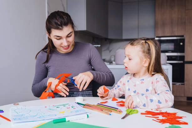 istock mother and 4-year-old daughter sit at white table in cozy modern kitchen, making crafts together. Child in heart-patterned pajamas focused on cutting paper hearts, mom gently helps creating. 2200372761