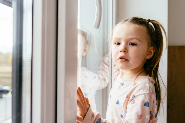 istock 4-year-old girl stands by window, looking outside with thoughtful expression. Her reflection in the glass adds depth to the moment. Dressed in heart-patterned pajamas, she seems lost in thought. 2200372781