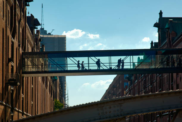 A modern bridge in Hamburg's Speicherstadt stands out against the bright sky, with people on the bridge appearing as silhouettes, creating a vibrant scene in an urban environment.